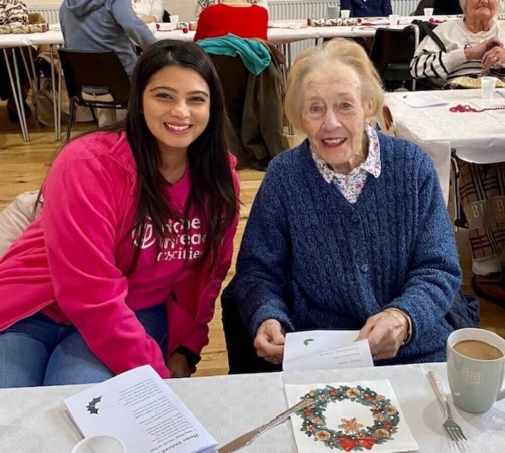 Two women sitting at a table with papers, smiling at the camera, one wearing a pink shirt and the other wearing a blue sweater. - Home Instead