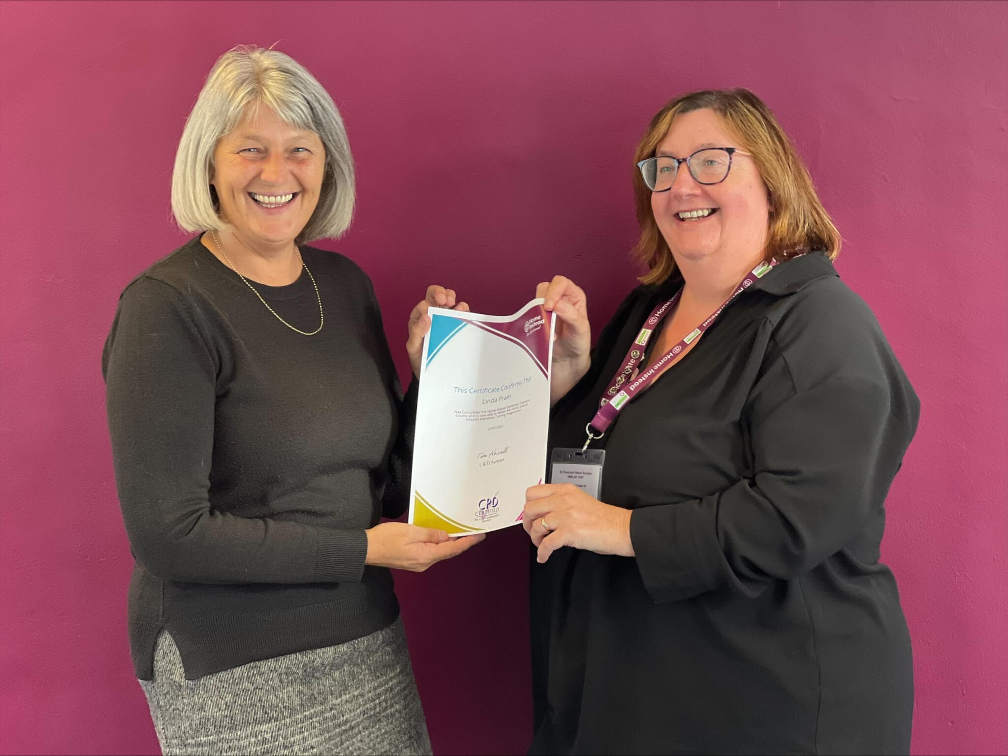 Two women smiling while holding a certificate against a purple wall background. - Home Instead