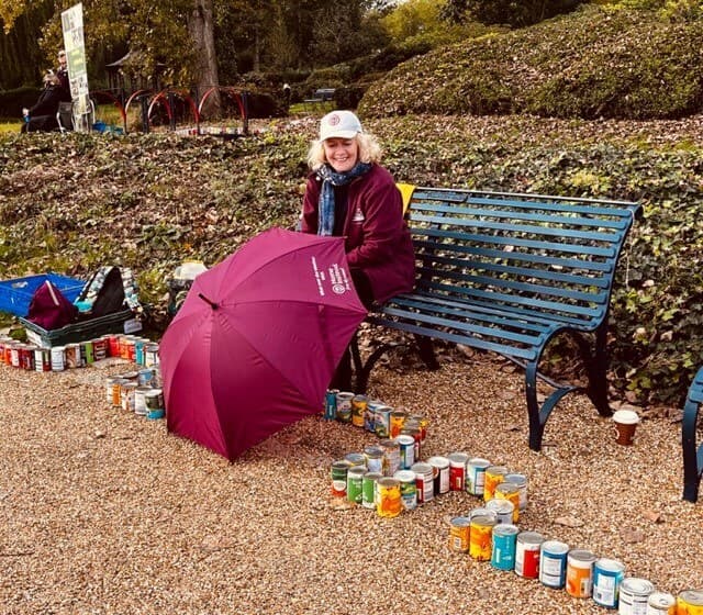 A person sitting on a bench outdoors with an umbrella and various canned goods arranged around. - Home Instead