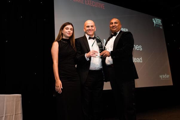 Three people in formal attire pose on stage with an award; a man in the middle holds the trophy, flanked by a woman and another man. - Home Instead