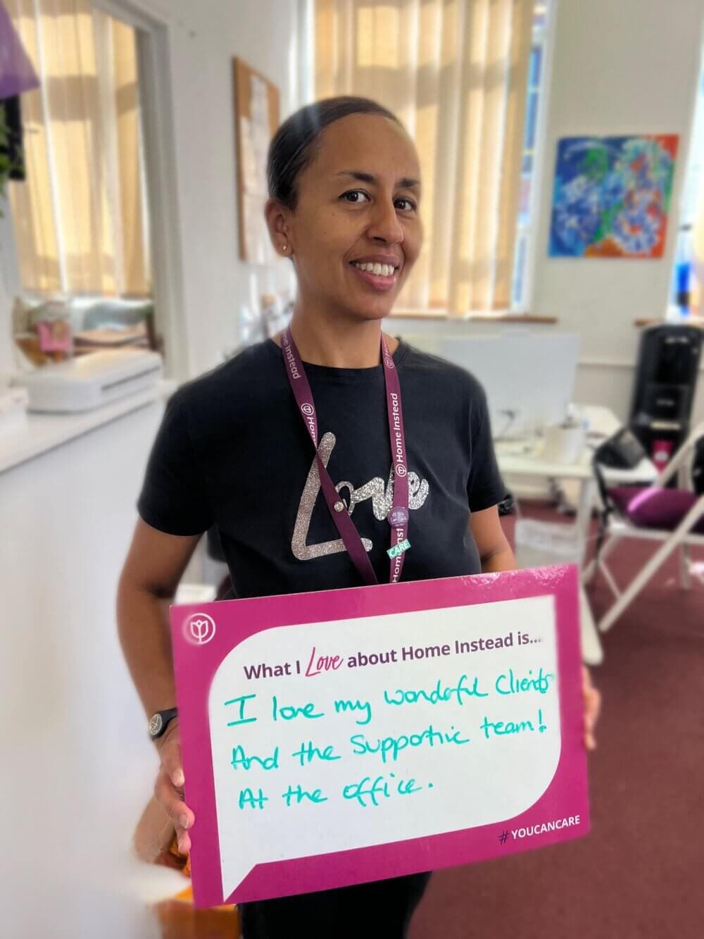 Person smiling and holding a sign that says they love their clients and supportive team at work, in an office setting. - Home Instead