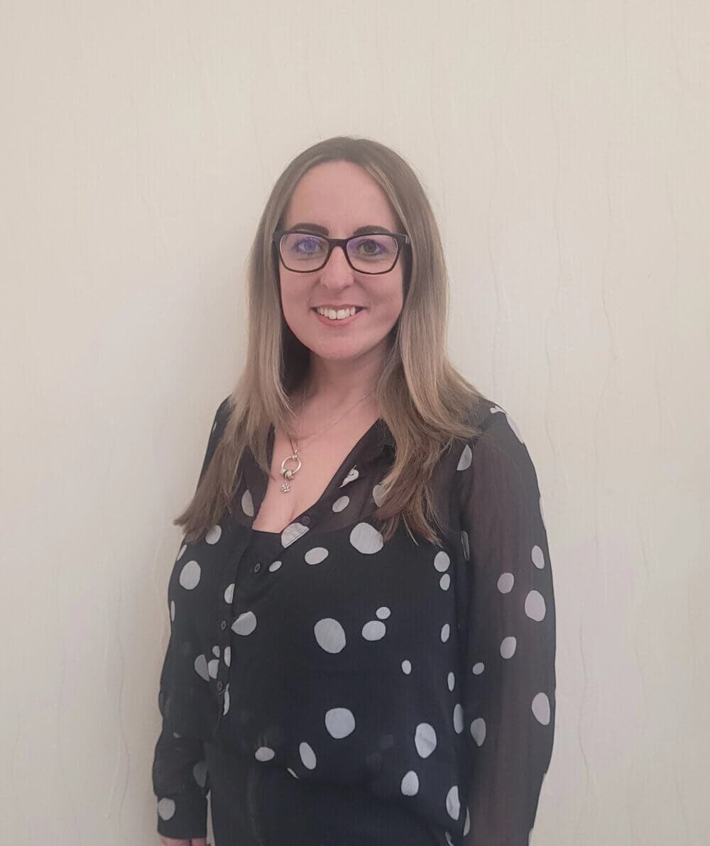 A woman with long hair and glasses, wearing a black blouse with white polka dots, smiles while standing against a plain wall. - Home Instead