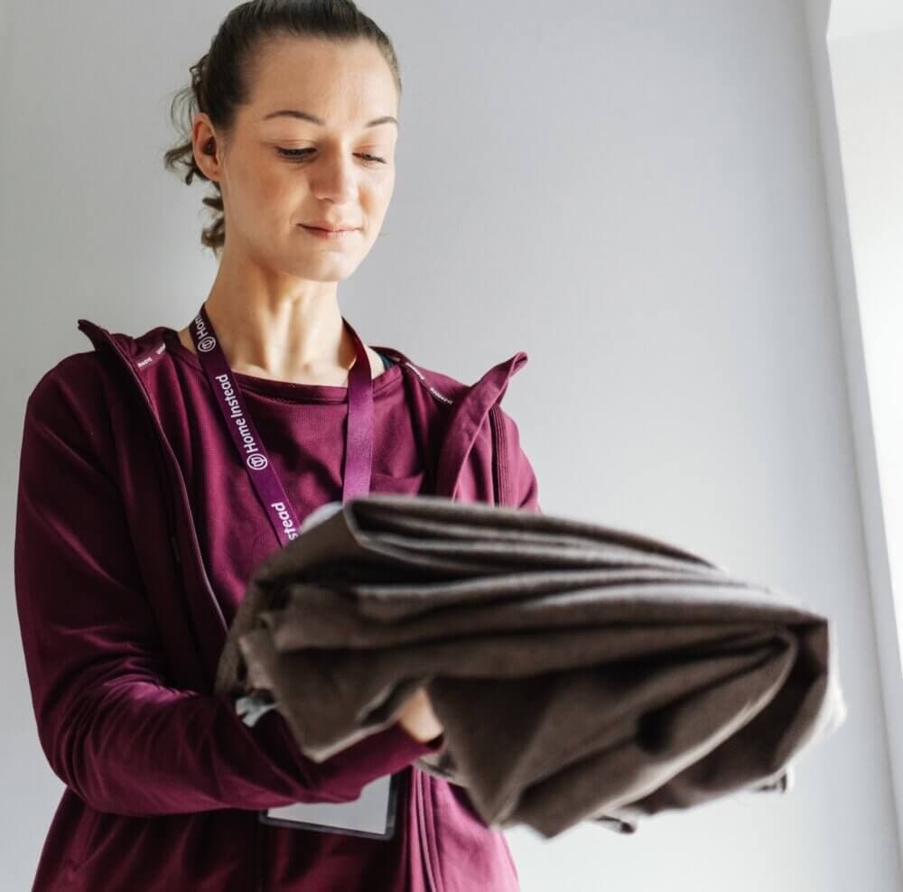 Person in maroon attire looking down while holding folded brown fabric, standing by a light-colored wall. - Home Instead