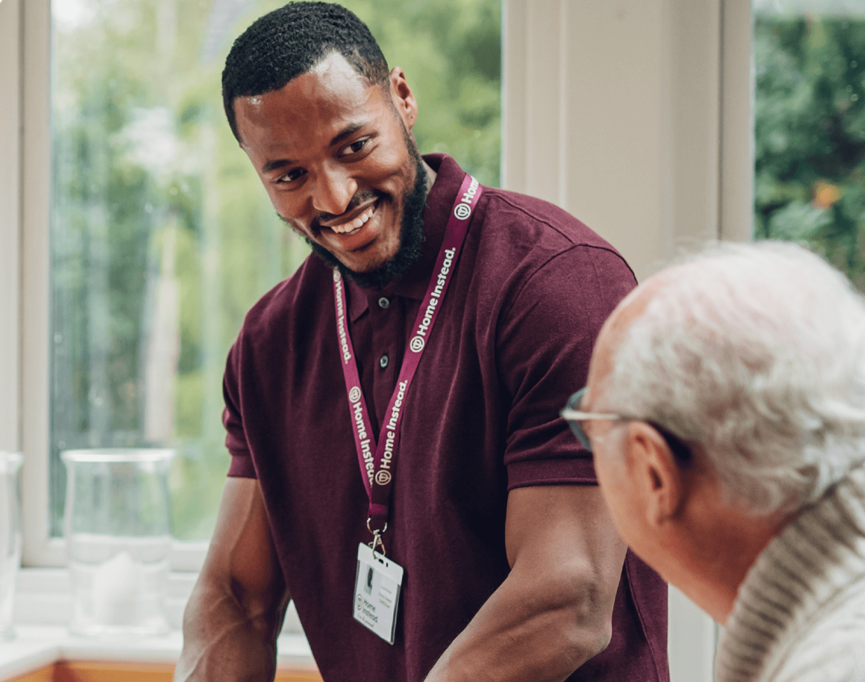 A man in a maroon shirt and lanyard smiles at an older man with glasses in a well-lit room. - Home Instead
