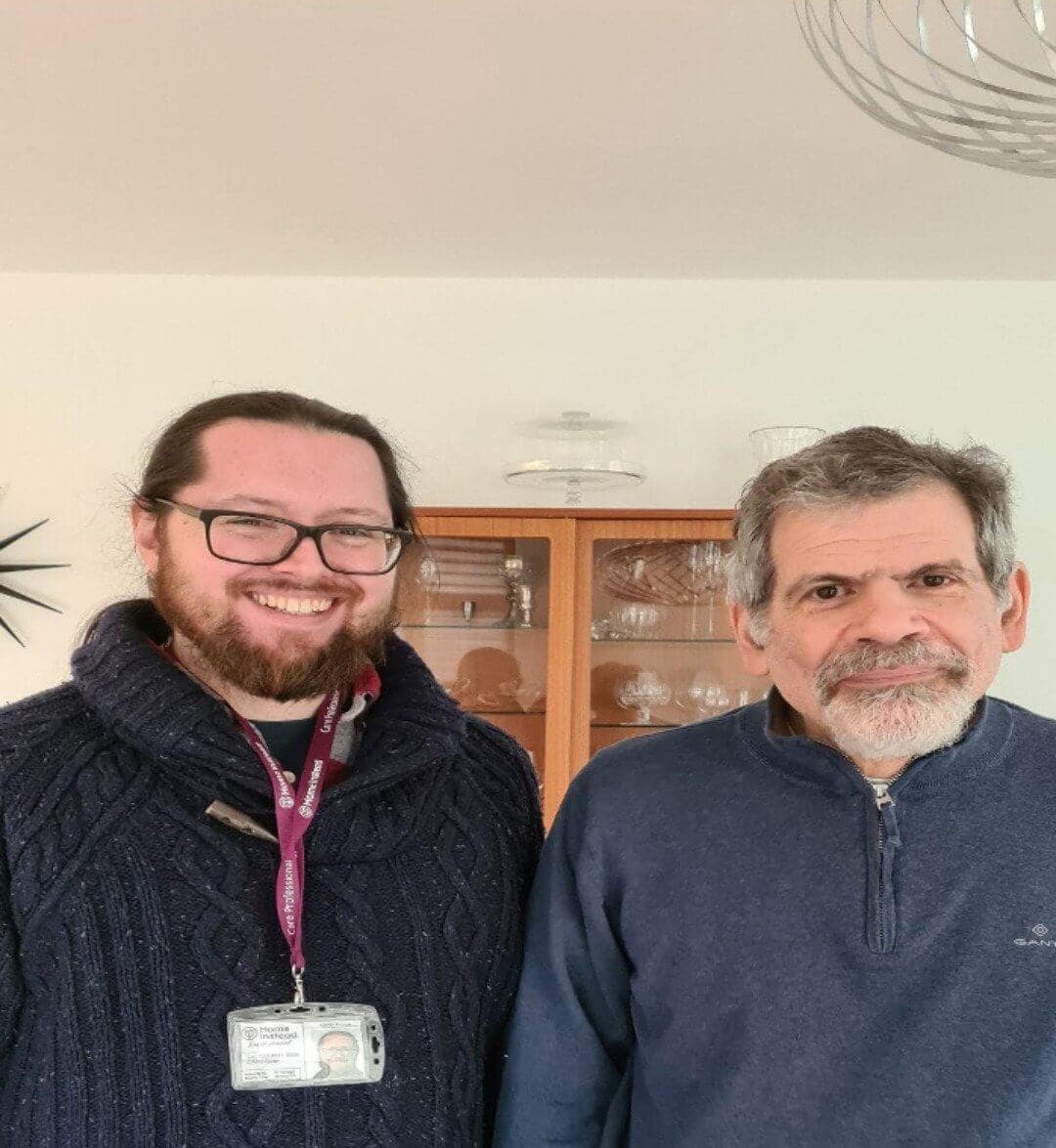 Two men are standing and smiling. One wears a lanyard with an ID badge, and both wear dark sweaters. - Home Instead