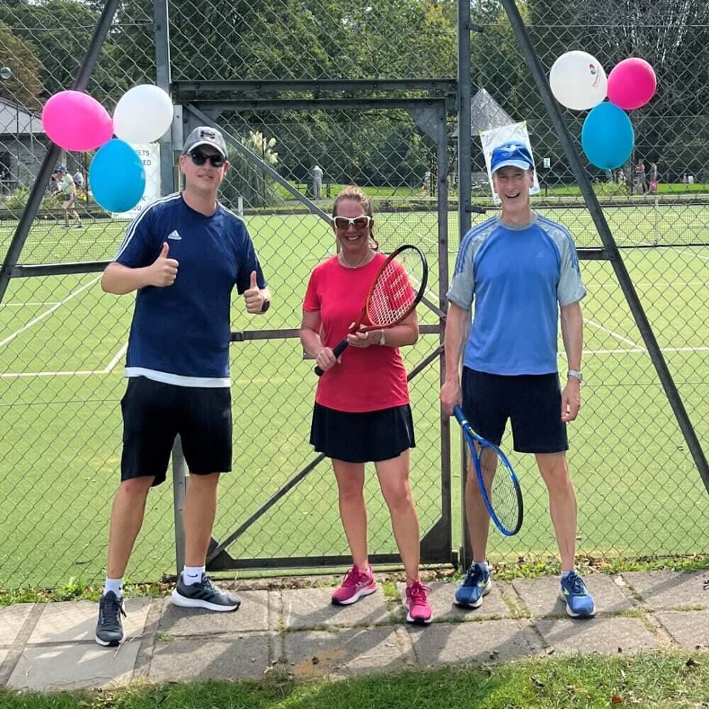 Three people are standing on a tennis court, holding rackets, with balloons attached to the fence behind them. - Home Instead
