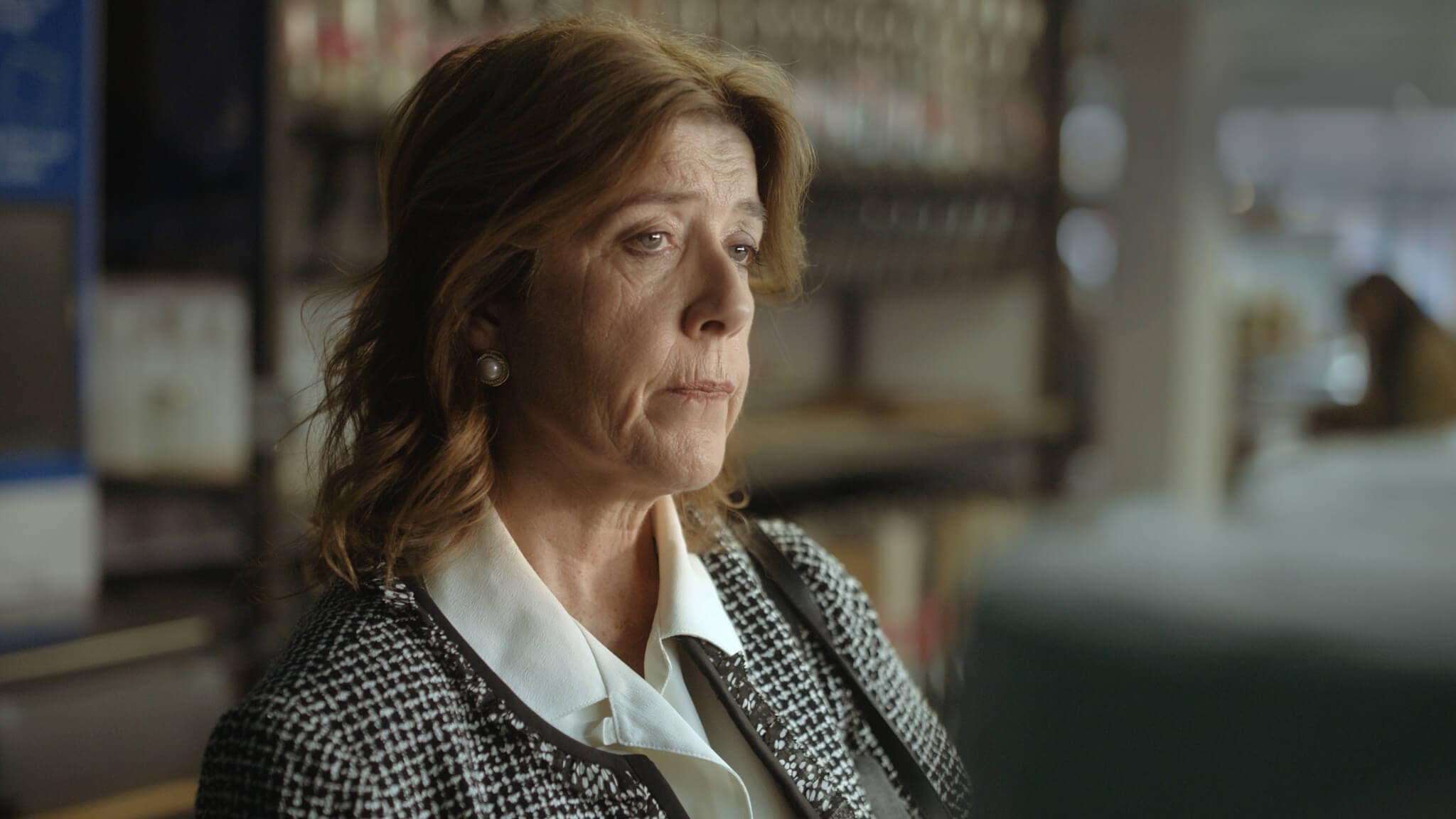 A woman with brown hair in a checkered blazer and white blouse looks pensive in an indoor setting. - Home Instead
