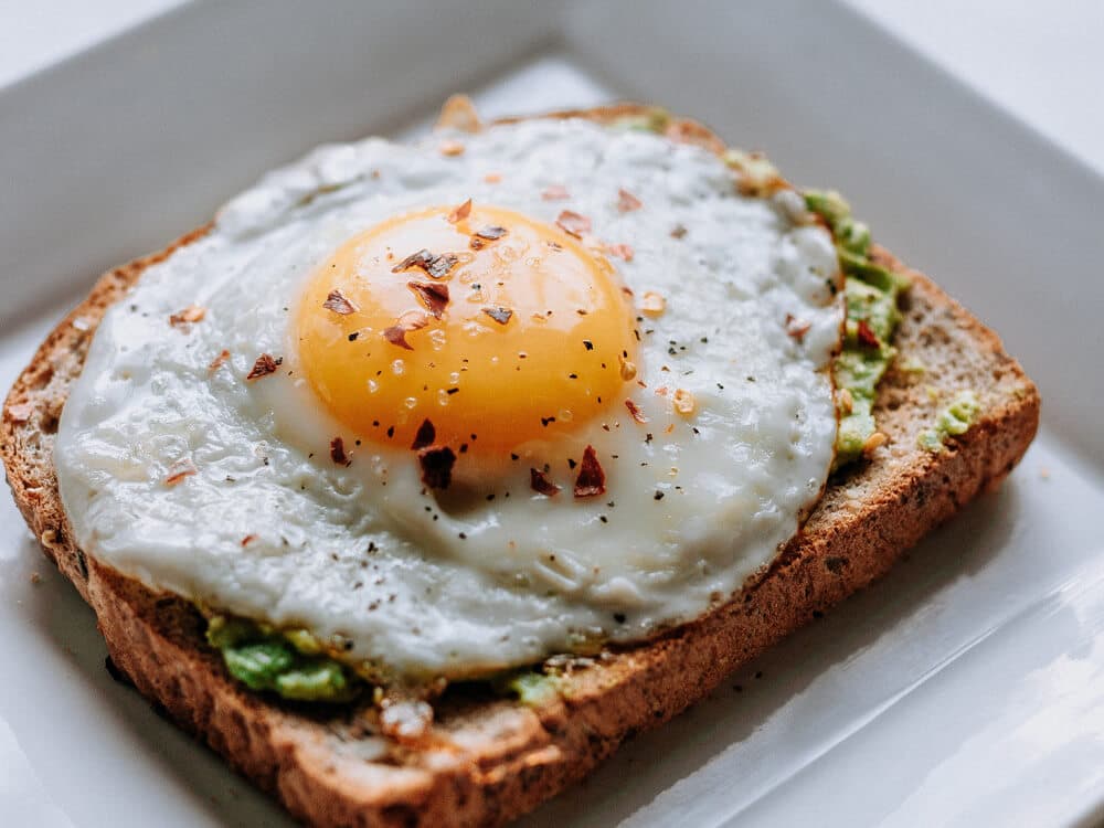A slice of toast topped with mashed avocado and a fried egg with a runny yolk, garnished with black pepper and chili flakes. - Home Instead