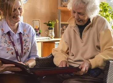 A young woman and an elderly woman smiling while looking at a photo album together in a cozy room. - Home Instead