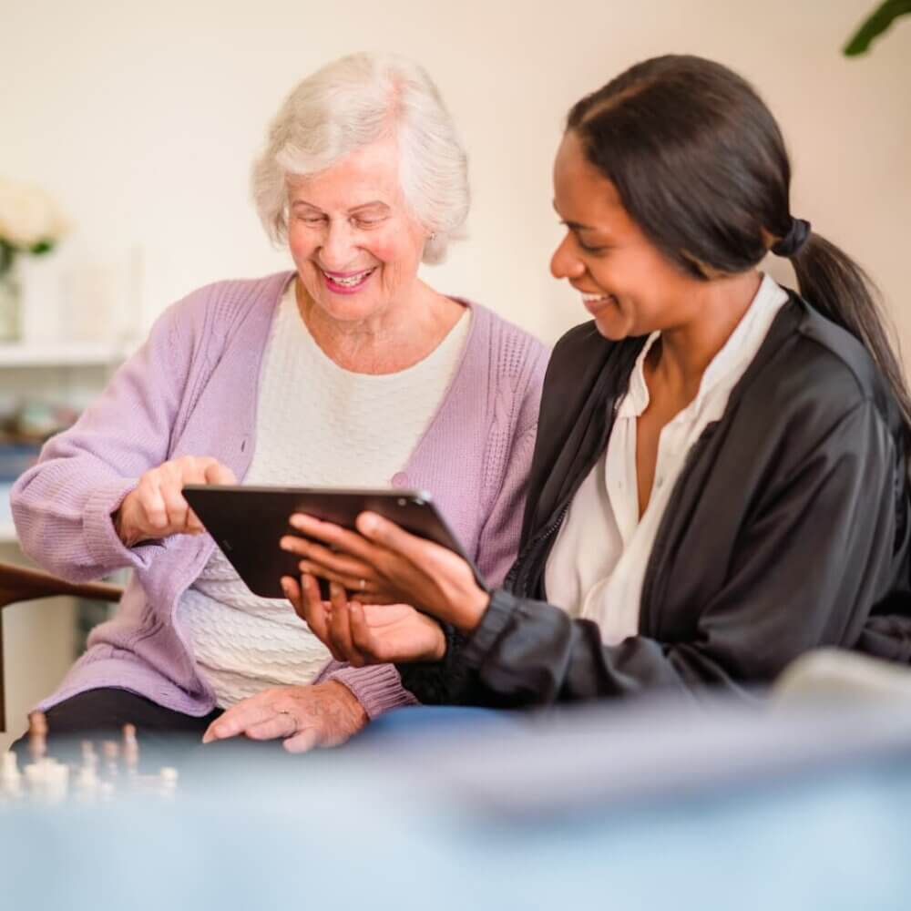 An elderly woman and a younger woman smiling while looking at a tablet together indoors. - Home Instead
