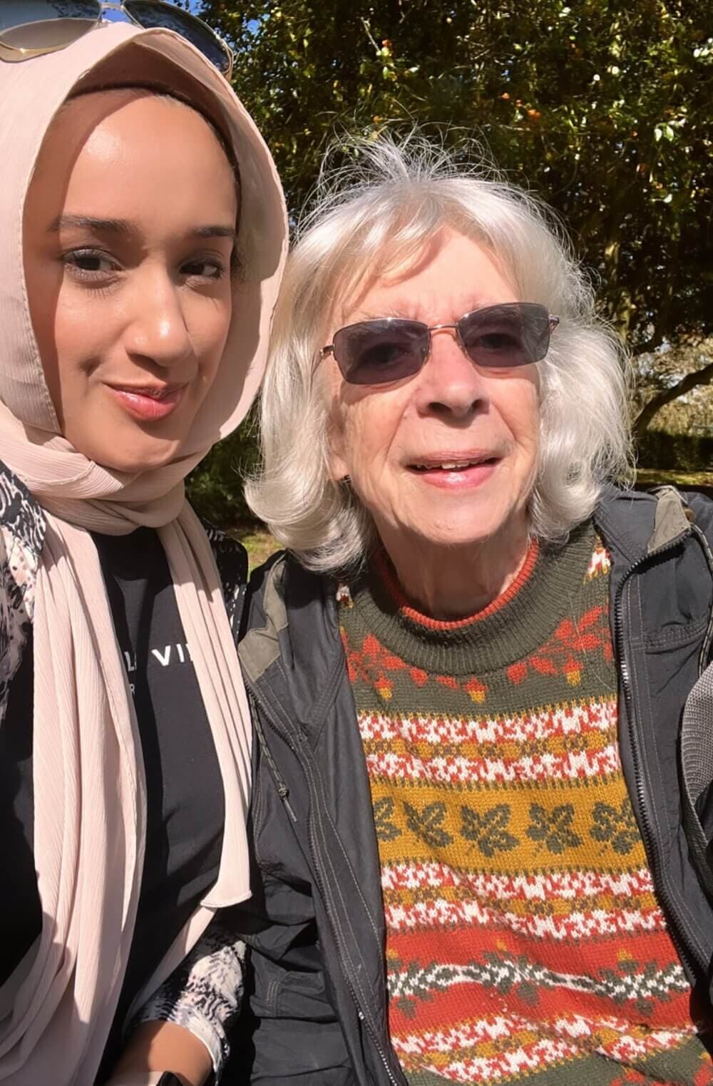 Two women smiling outdoors, one wearing a hijab and the other wearing sunglasses and a festive sweater. - Home Instead