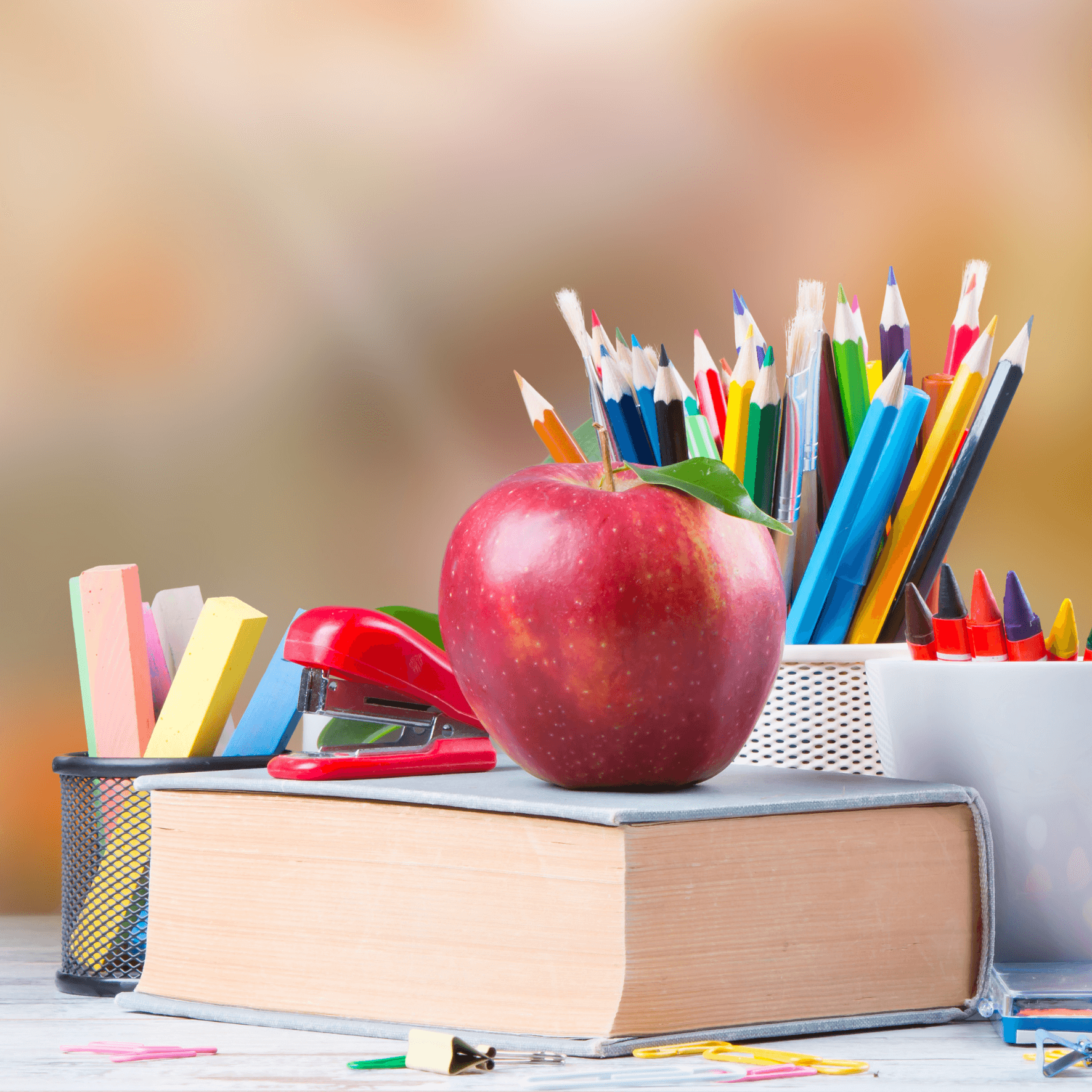 A red apple on a stack of books next to colorful pencils, pens, and a red stapler on a desk, depicting kids and family life. - Home Instead