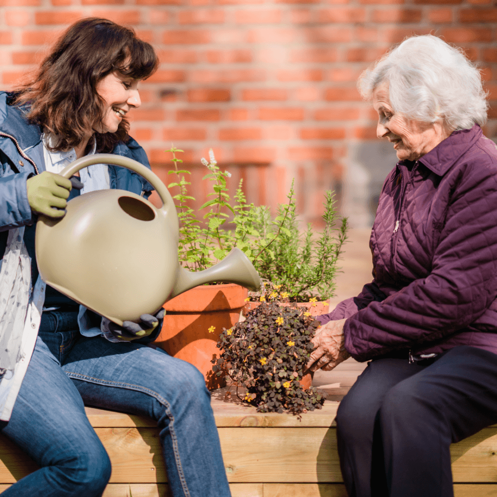 Two women gardening together, one holding a watering can and the other tending to plants in pots, with a brick wall backdrop. - Home Instead