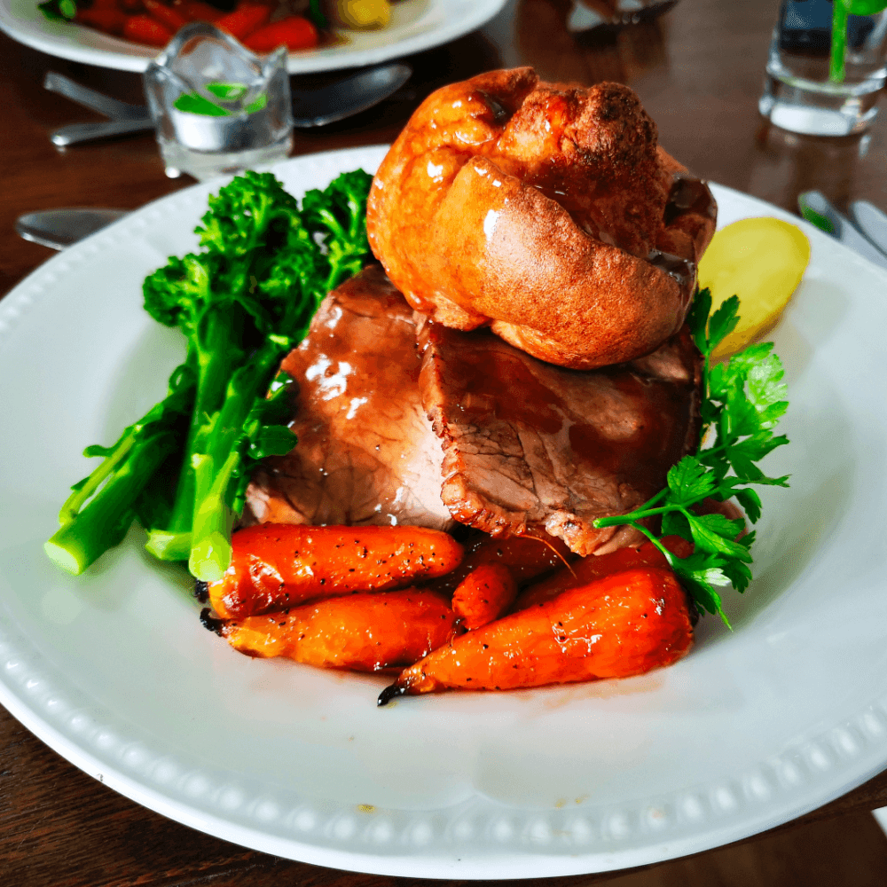 A plate of roast beef with gravy, Yorkshire pudding, glazed carrots, broccoli, and a potato slice. - Home Instead
