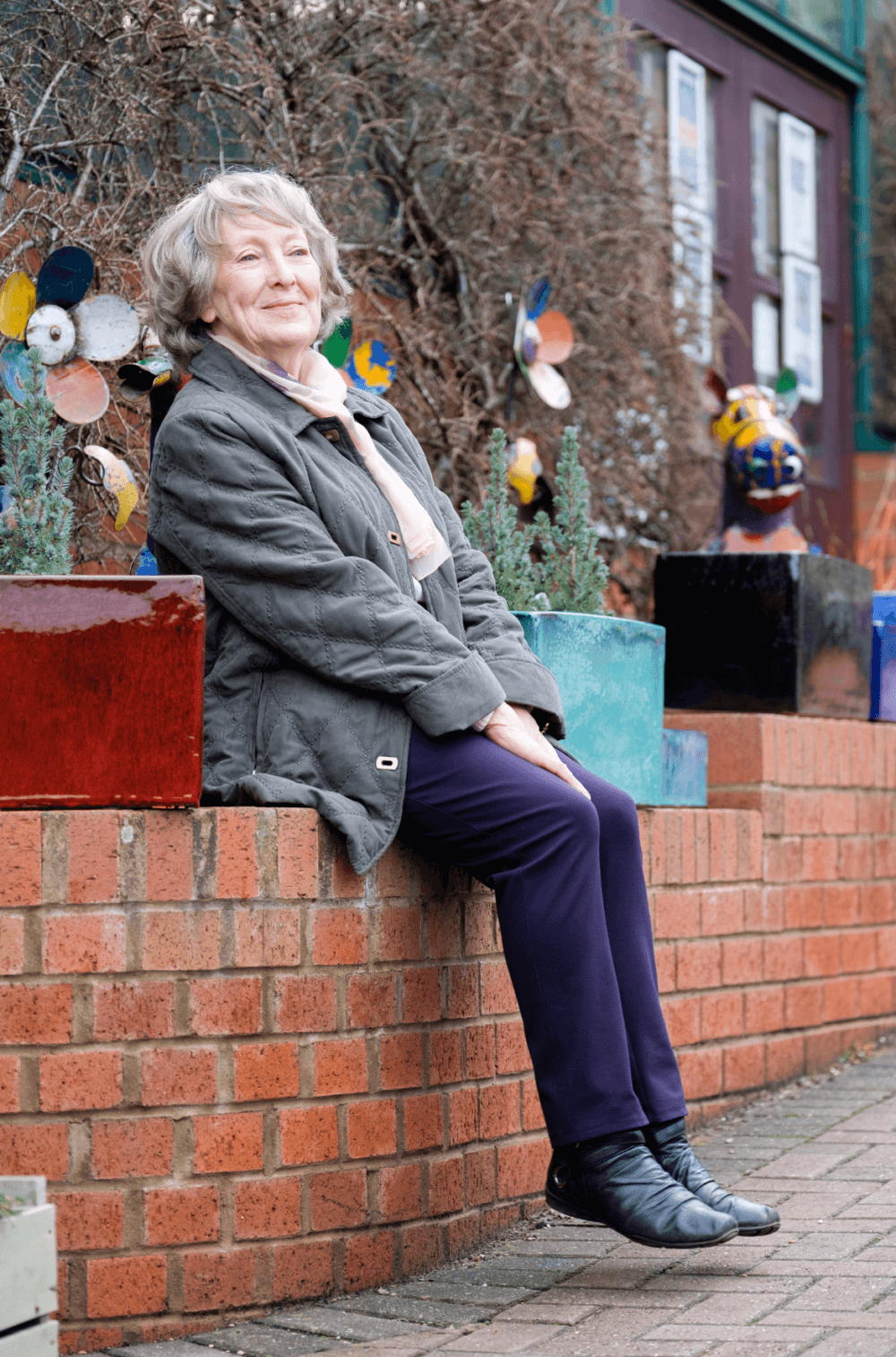 Elderly woman with gray hair and winter clothing sitting on a brick wall, decorated with colorful sculptures. - Home Instead