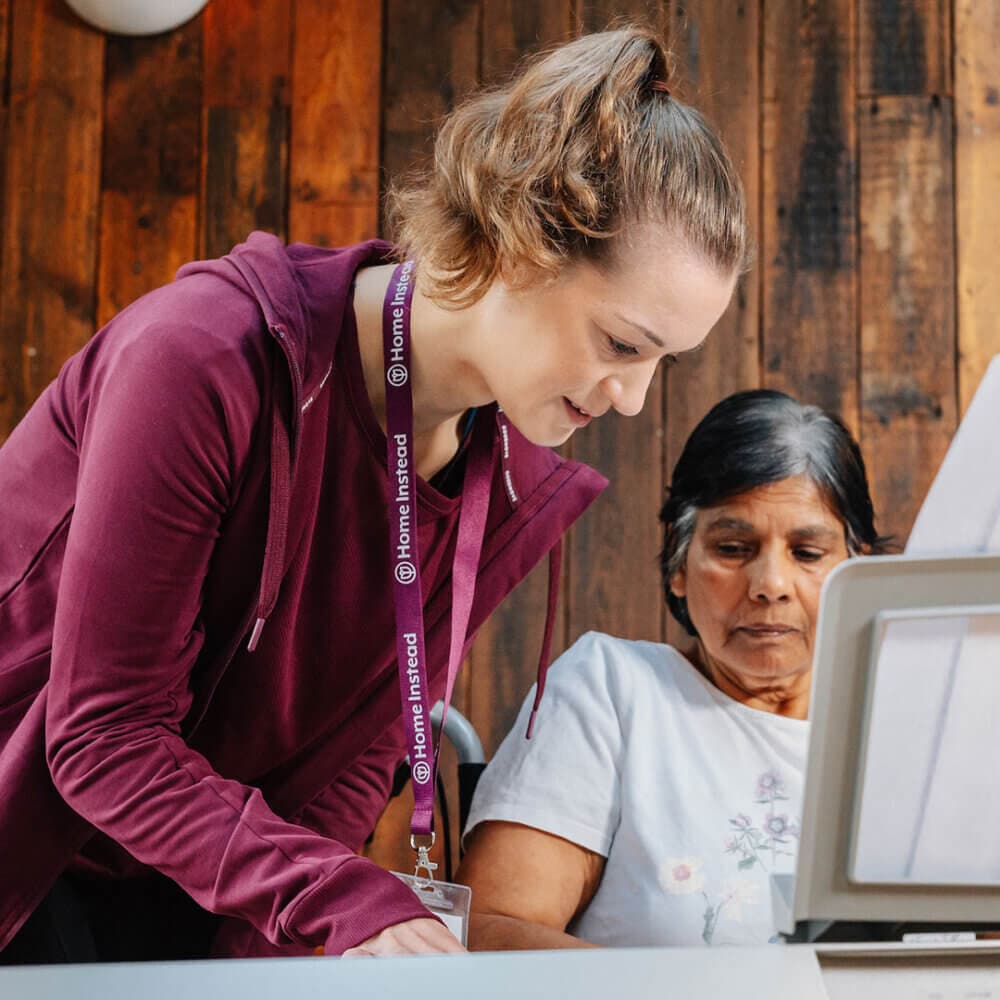 A woman in a maroon jacket assists an older woman using a laptop at a wooden table. - Home Instead