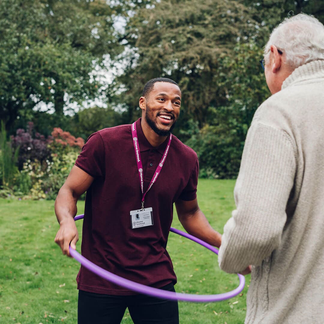 A man with a name badge helps an elderly man use a hula hoop outside in a garden. - Home Instead