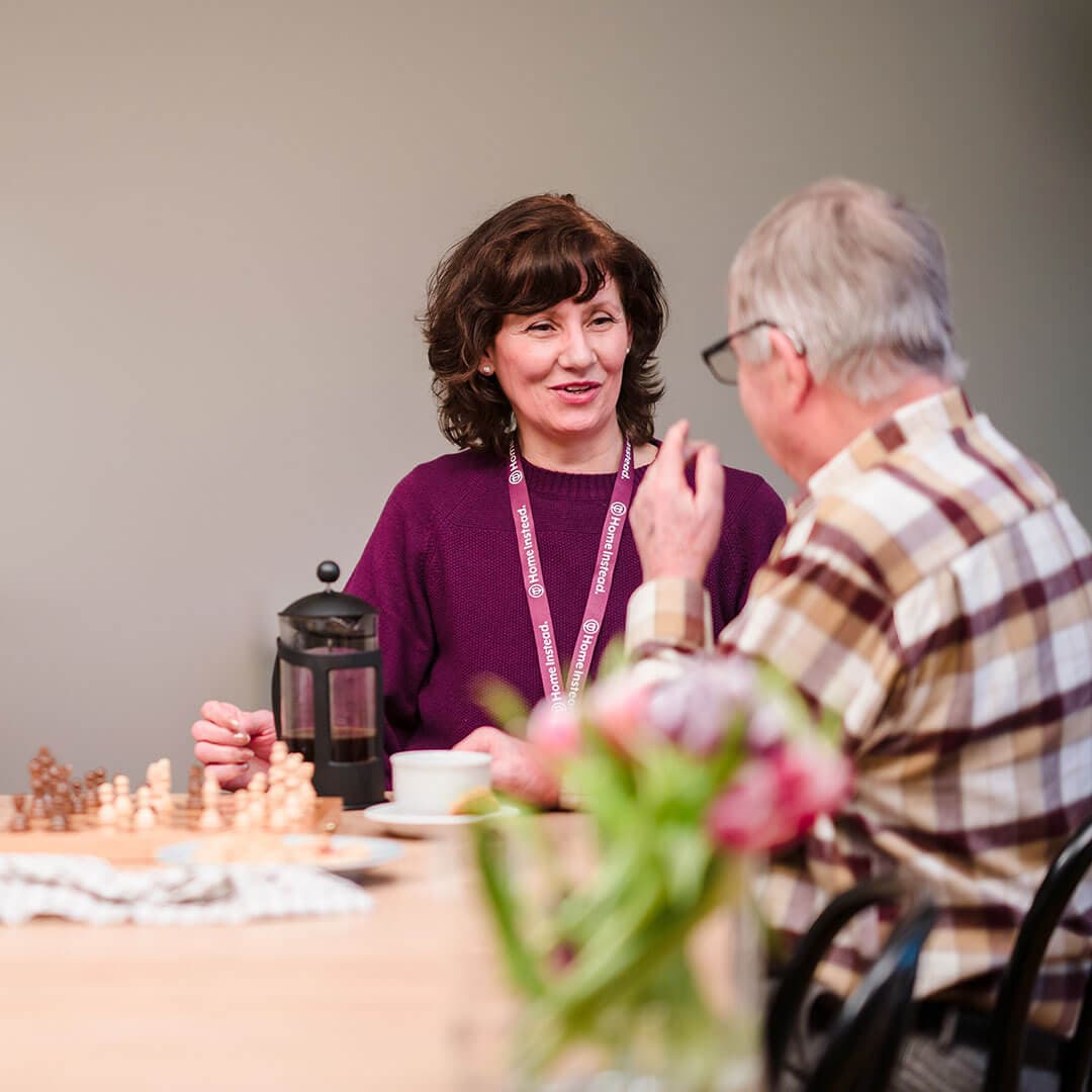 A woman and an older man sitting at a table with a chessboard, flowers, and a French press, engaged in conversation. - Home Instead