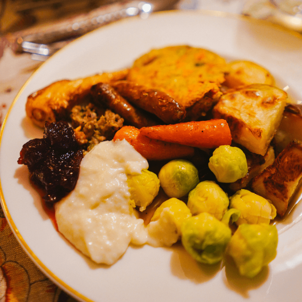 A plate of food featuring vegetables, sausages, mashed potatoes, a dollop of sauce, and roasted root vegetables. - Home Instead
