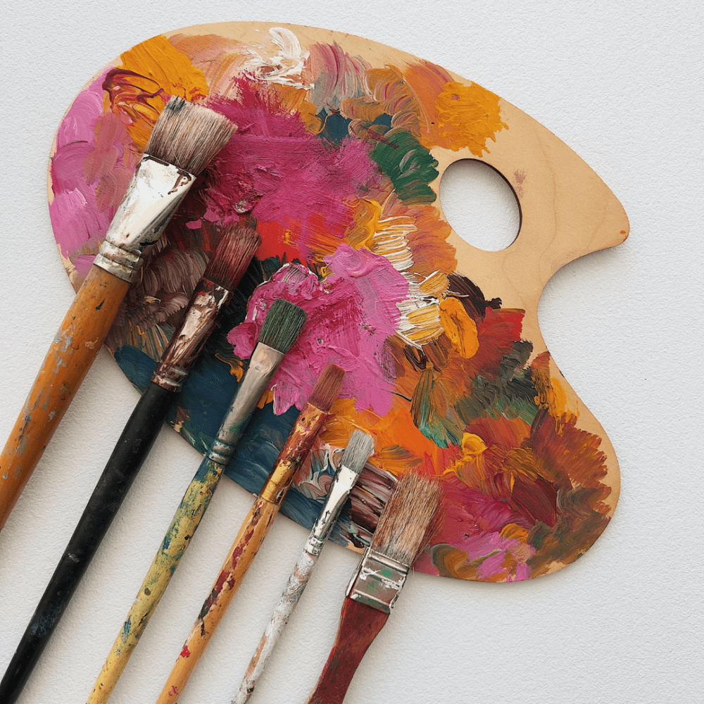 A wooden artist's palette with vibrant paint and six paintbrushes arranged on a light background. - Home Instead