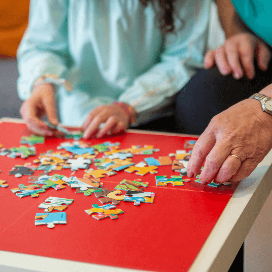 Two people assembling a colorful jigsaw puzzle on a red table, their hands working together on the pieces. - Home Instead