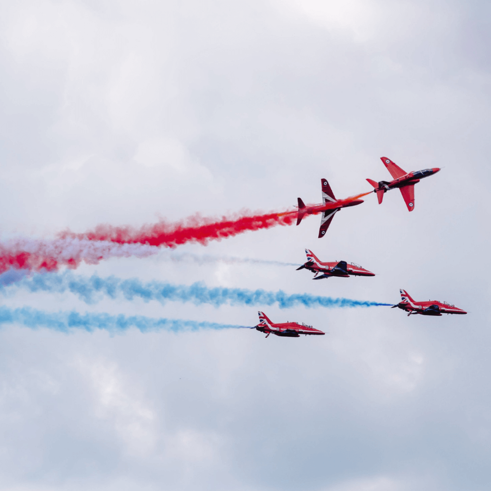 Five red jets perform an aerial show with red, white, and blue smoke trails against a cloudy sky. - Home Instead