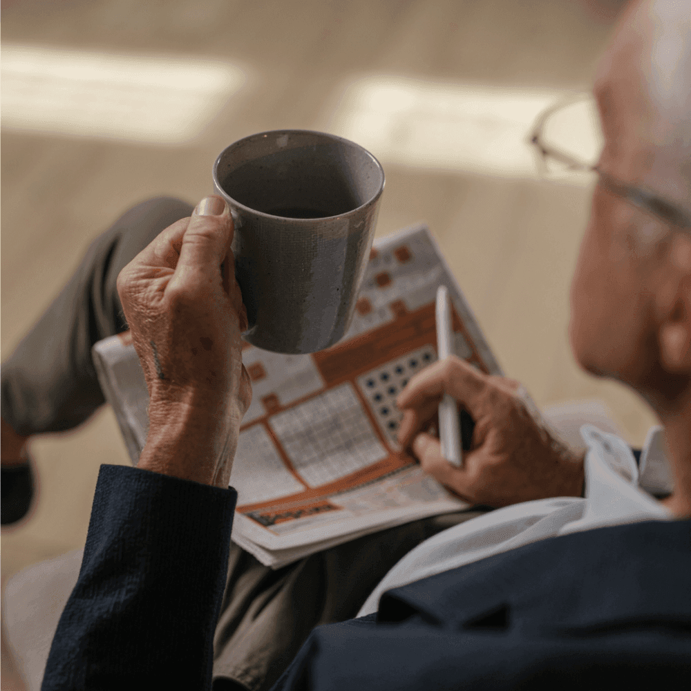 A person holding a mug and pen while working on a crossword puzzle in a newspaper. - Home Instead