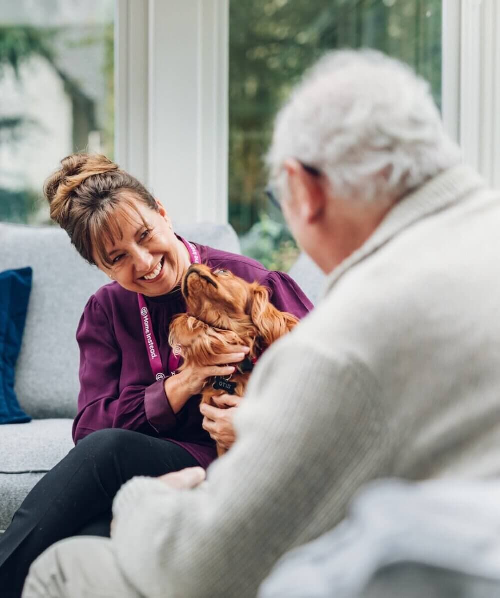 Woman smiling while holding a dog, sitting on a couch with an elderly man in a white sweater. - Home Instead