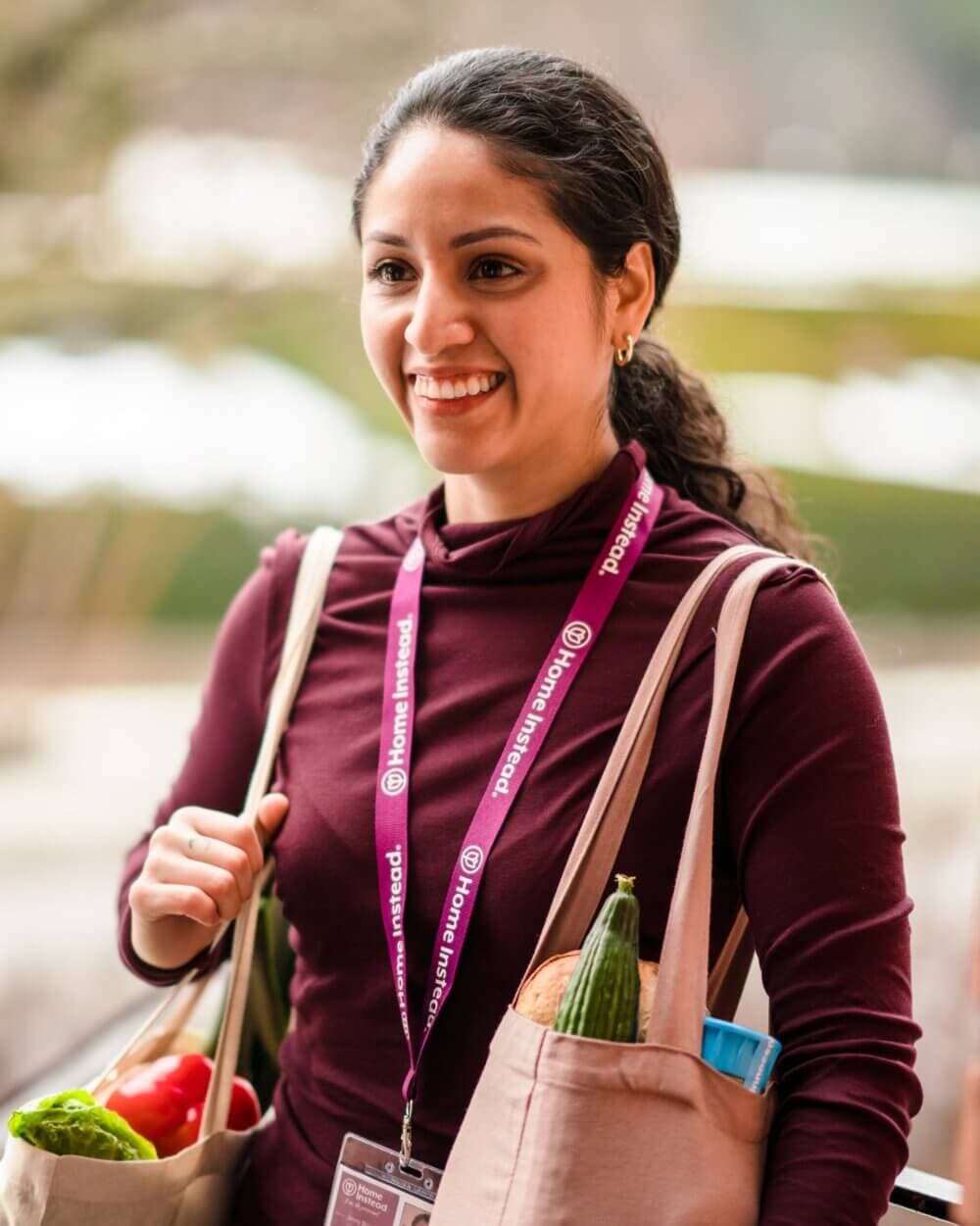A smiling woman carrying grocery bags, wearing a maroon top and a lanyard that reads "Home Instead. - Home Instead