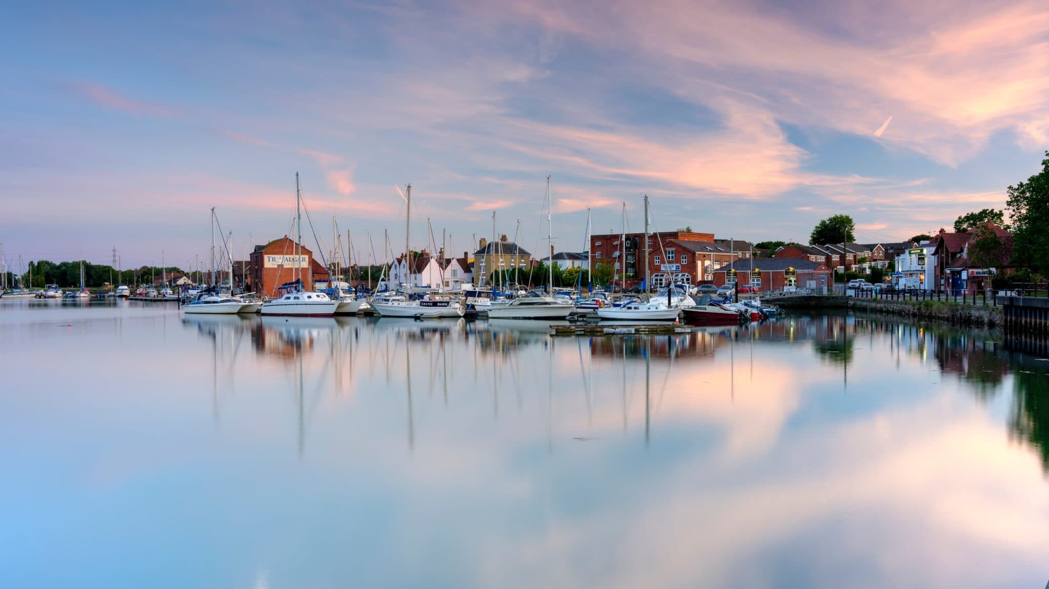 A serene harbor scene at sunset, with boats docked near buildings and calm water reflecting the sky and surrounding structures. - Home Instead