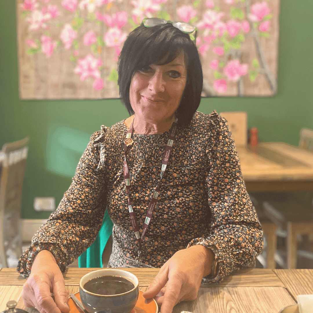 Woman sitting at a wooden table in a cafe, smiling and holding a cup of coffee, with floral artwork on the green wall behind. - Home Instead