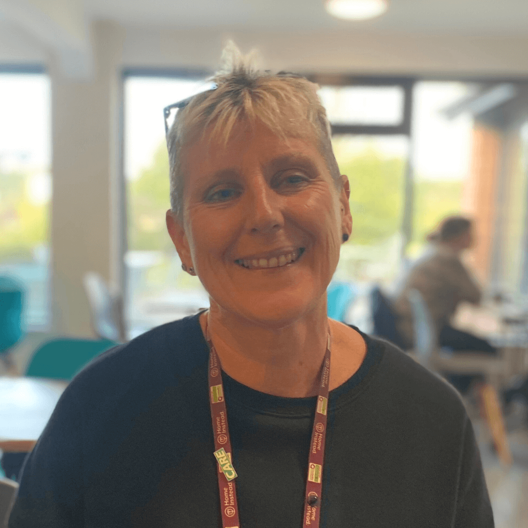 Smiling person with short hair wearing glasses and a lanyard, seated in a brightly lit room with people in the background. - Home Instead