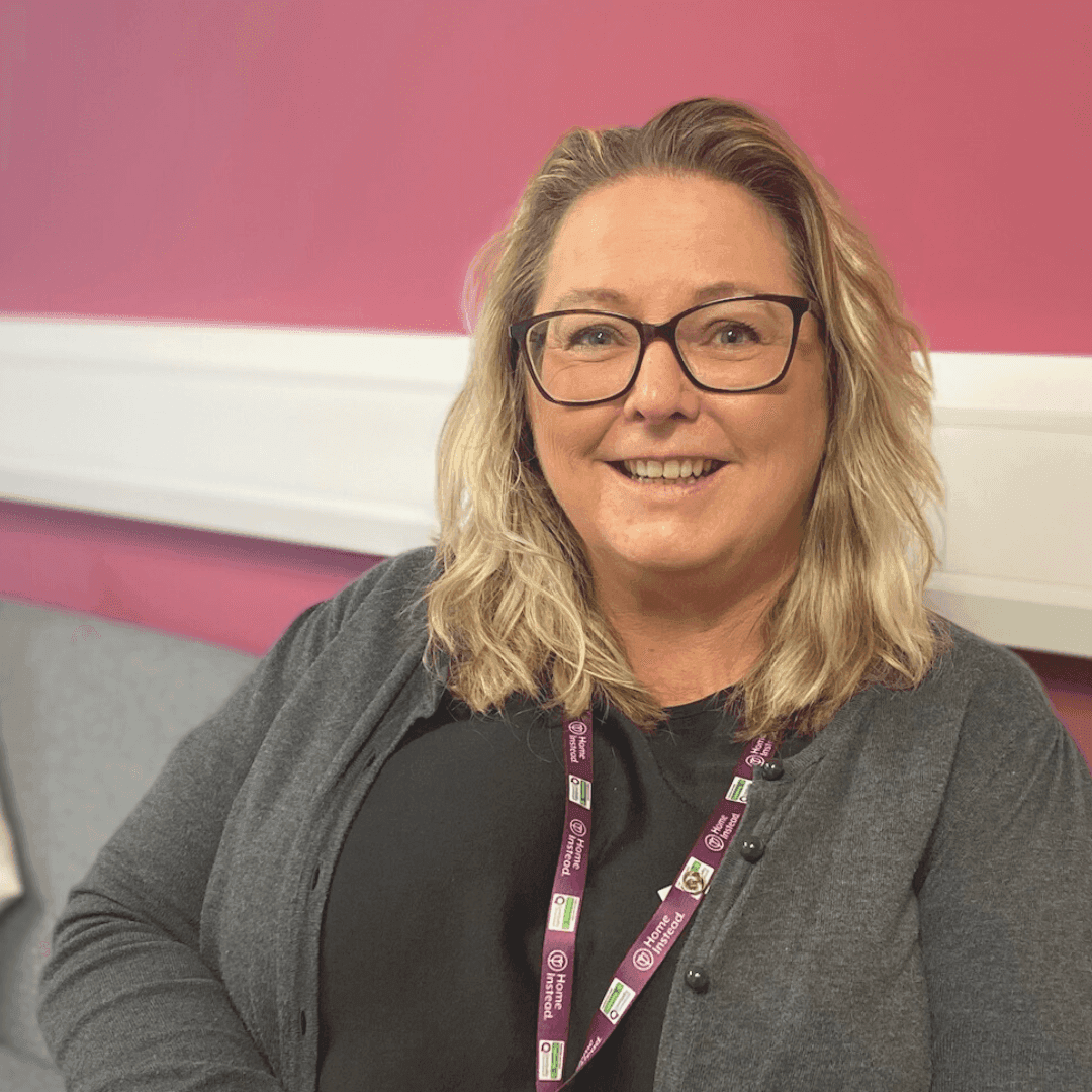 Smiling person with long blonde hair and glasses wearing a grey cardigan and lanyard, seated against a pink wall. - Home Instead
