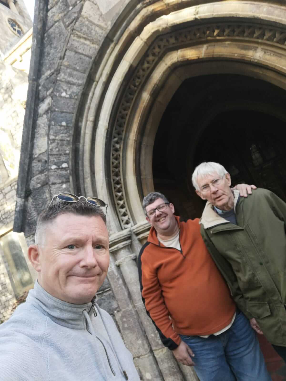 Three men standing in front of a stone archway, smiling at the camera. One is taking a selfie. - Home Instead