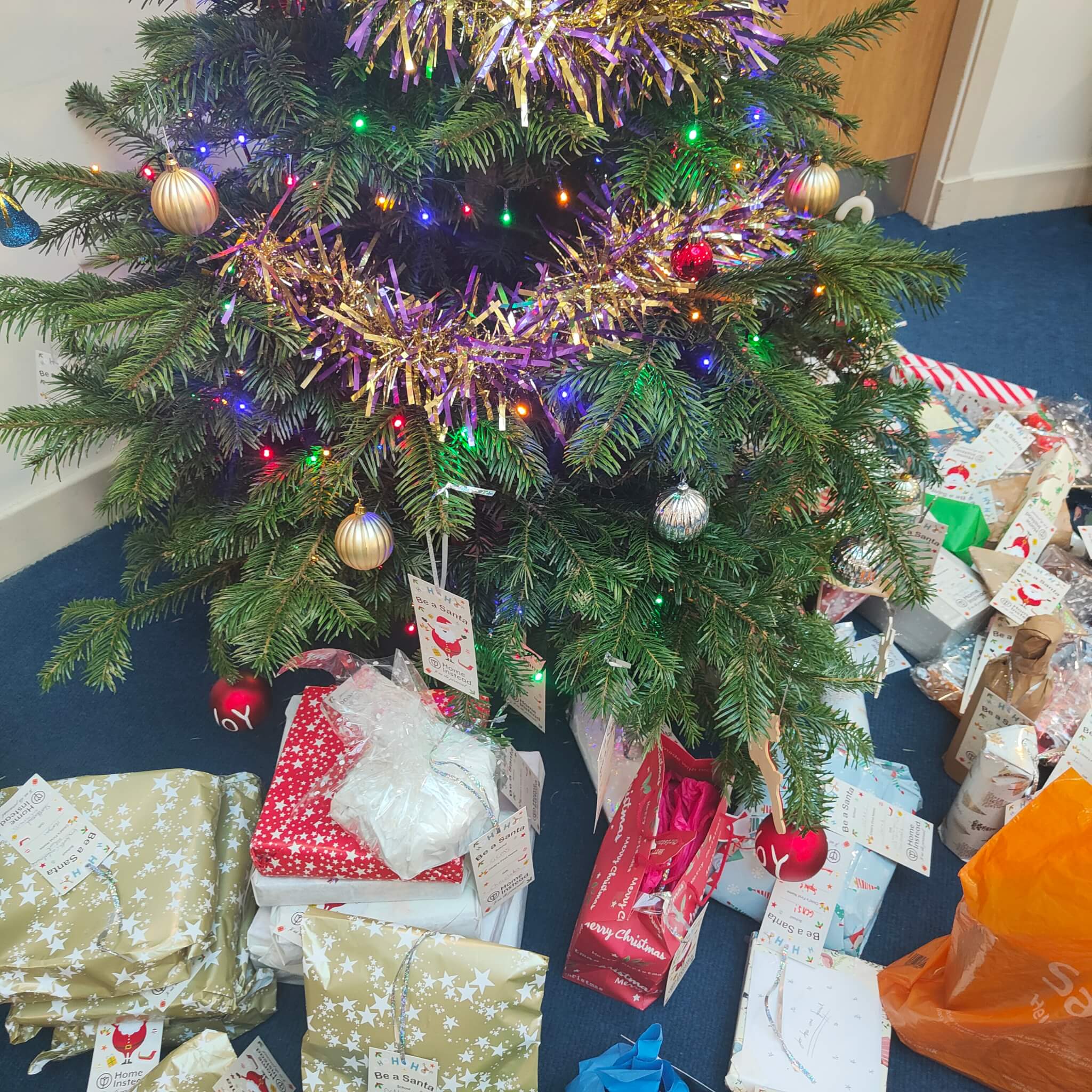 A decorated Christmas tree with colorful lights and tinsel, surrounded by numerous wrapped presents underneath. - Home Instead
