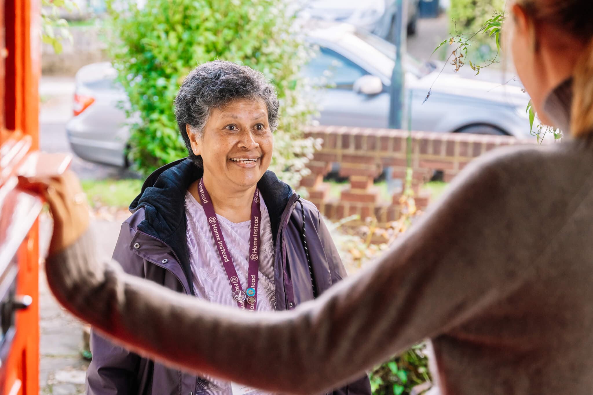 A smiling woman stands at a doorstep, wearing a purple lanyard, while another person opens the door to greet her. - Home Instead