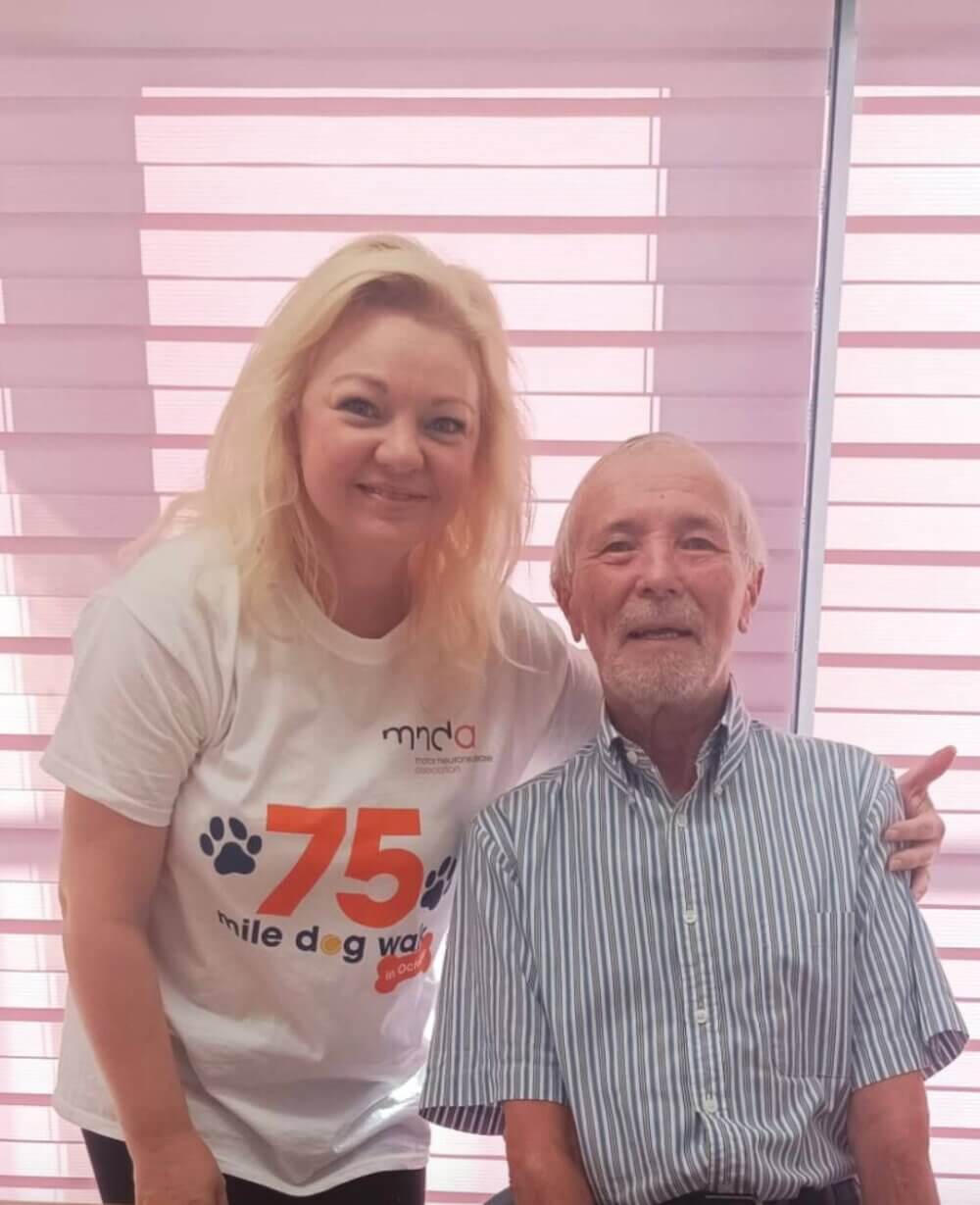 A woman in a white shirt and an elderly man in a striped shirt smile in front of a bright window. - Home Instead