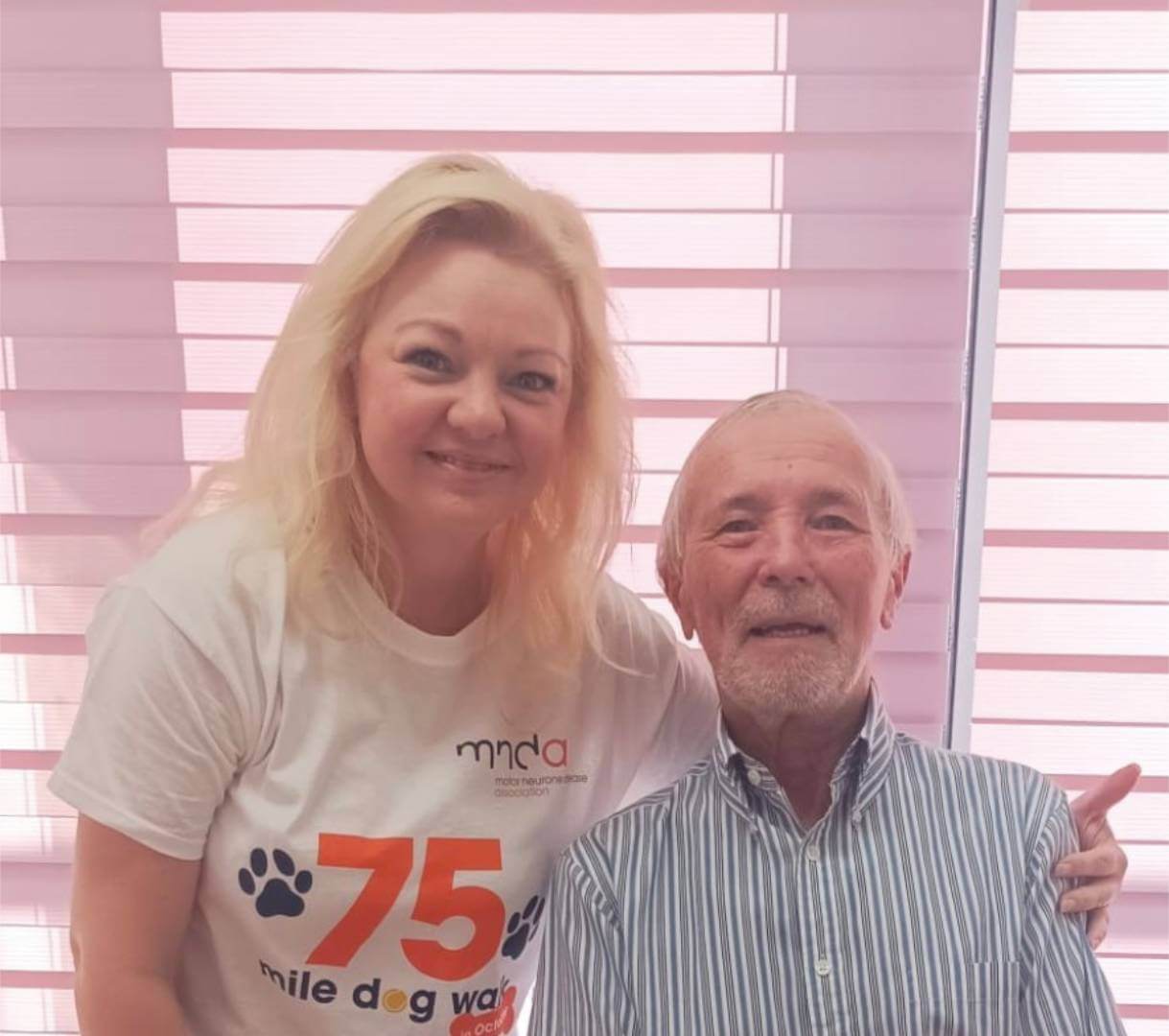 Two people smiling, a woman in a white t-shirt with "75 mile dog walk" logo and an elderly man in a striped shirt, in front of blinds. - Home Instead