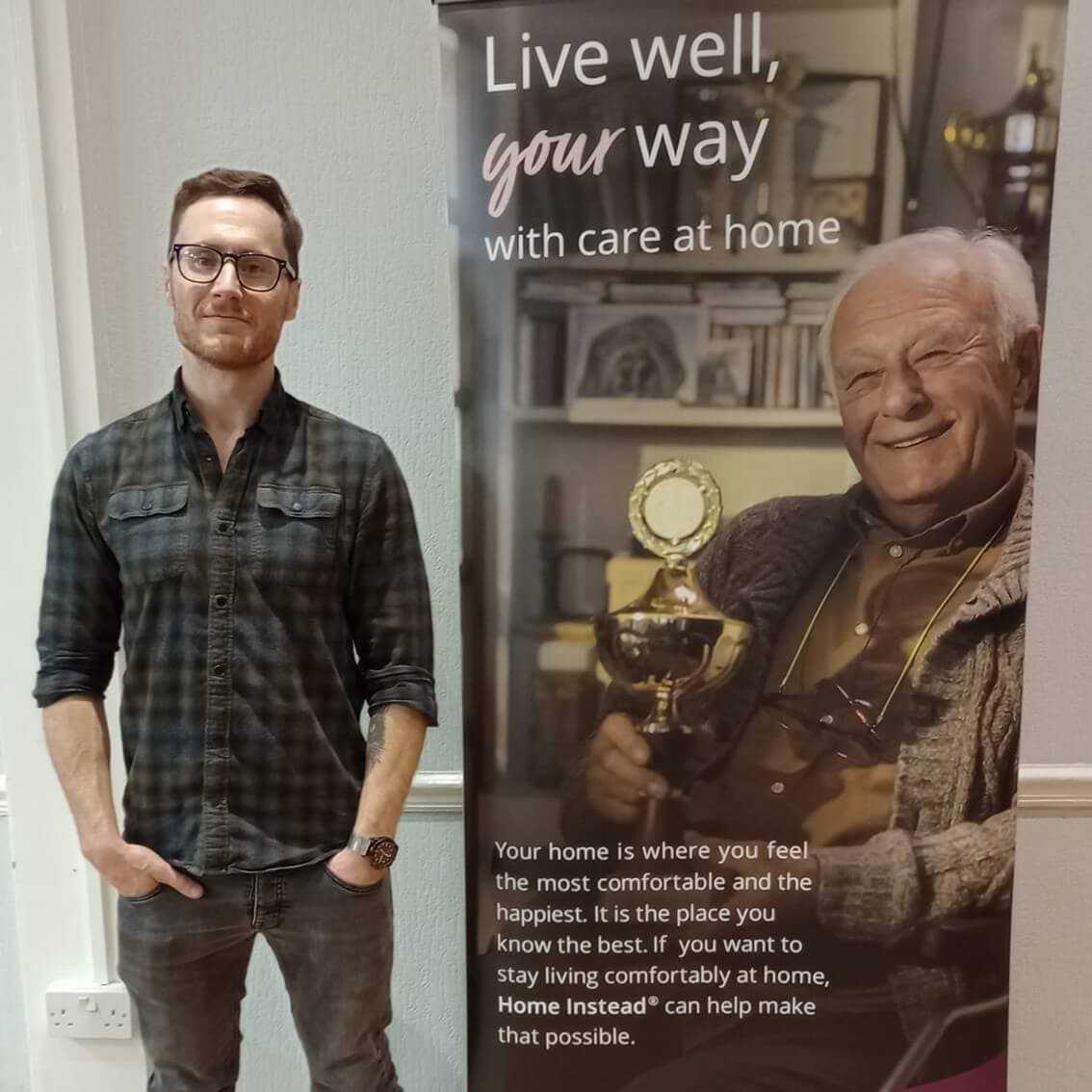 A man stands beside a banner showcasing an elderly man with a trophy, promoting Home Instead® care services. - Home Instead
