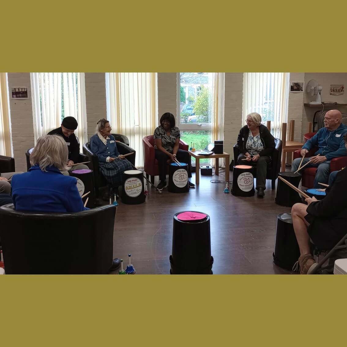 A group of people sitting in a circle in a room, each holding drumsticks and seated on bucket-like drums. - Home Instead