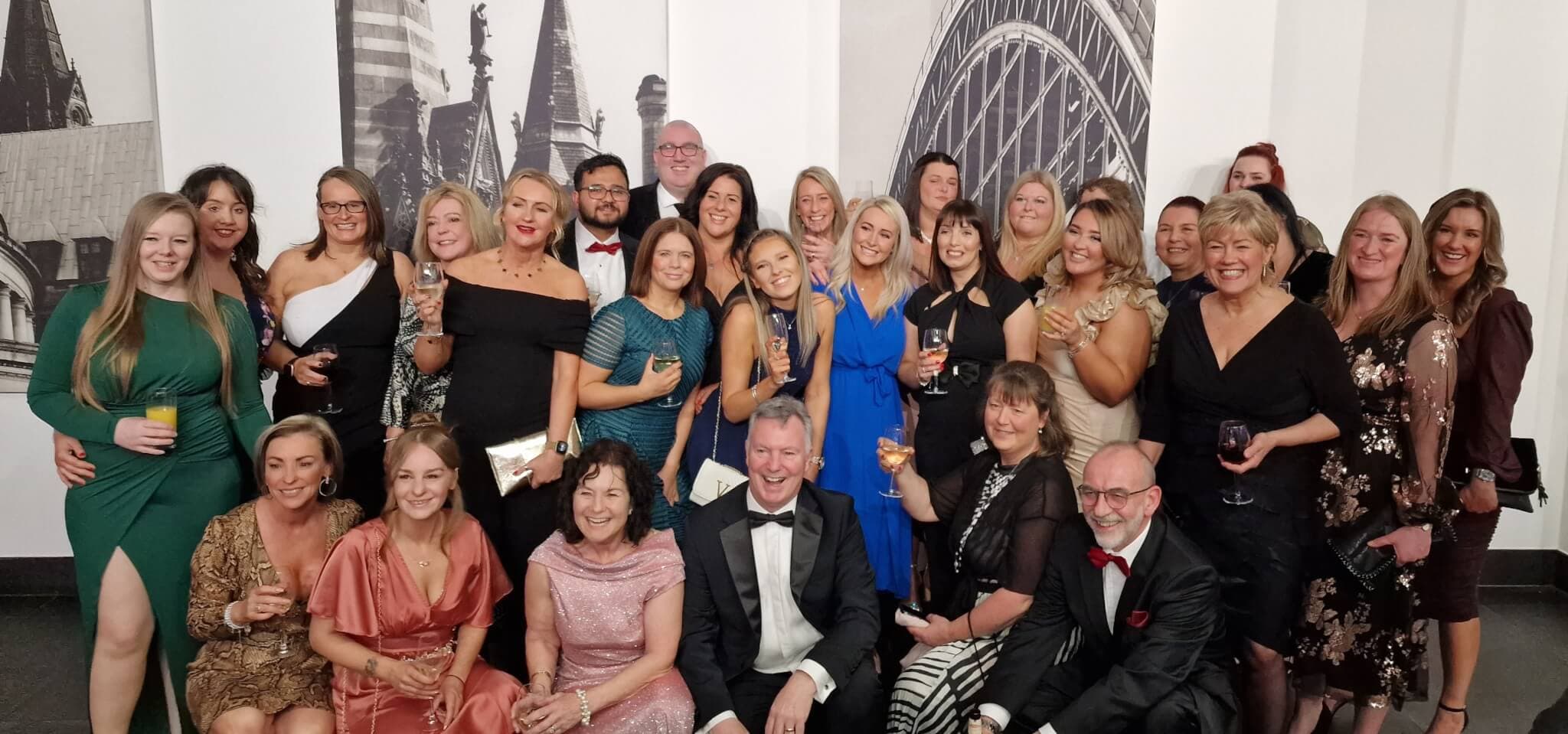 A group of people in formal attire smiling and holding drinks, posing for a group photo in front of a cityscape background. - Home Instead