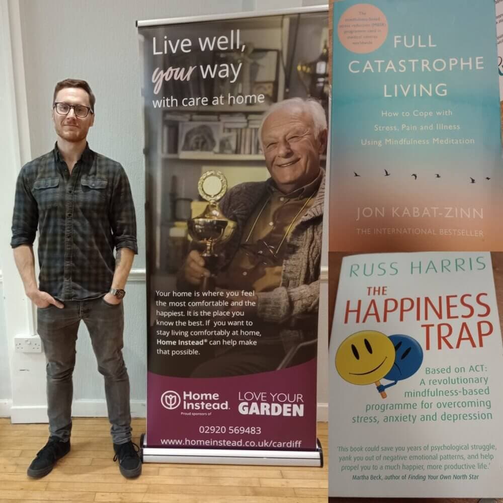 A man standing beside banners for Home Instead, "Full Catastrophe Living," and "The Happiness Trap" at an event. - Home Instead