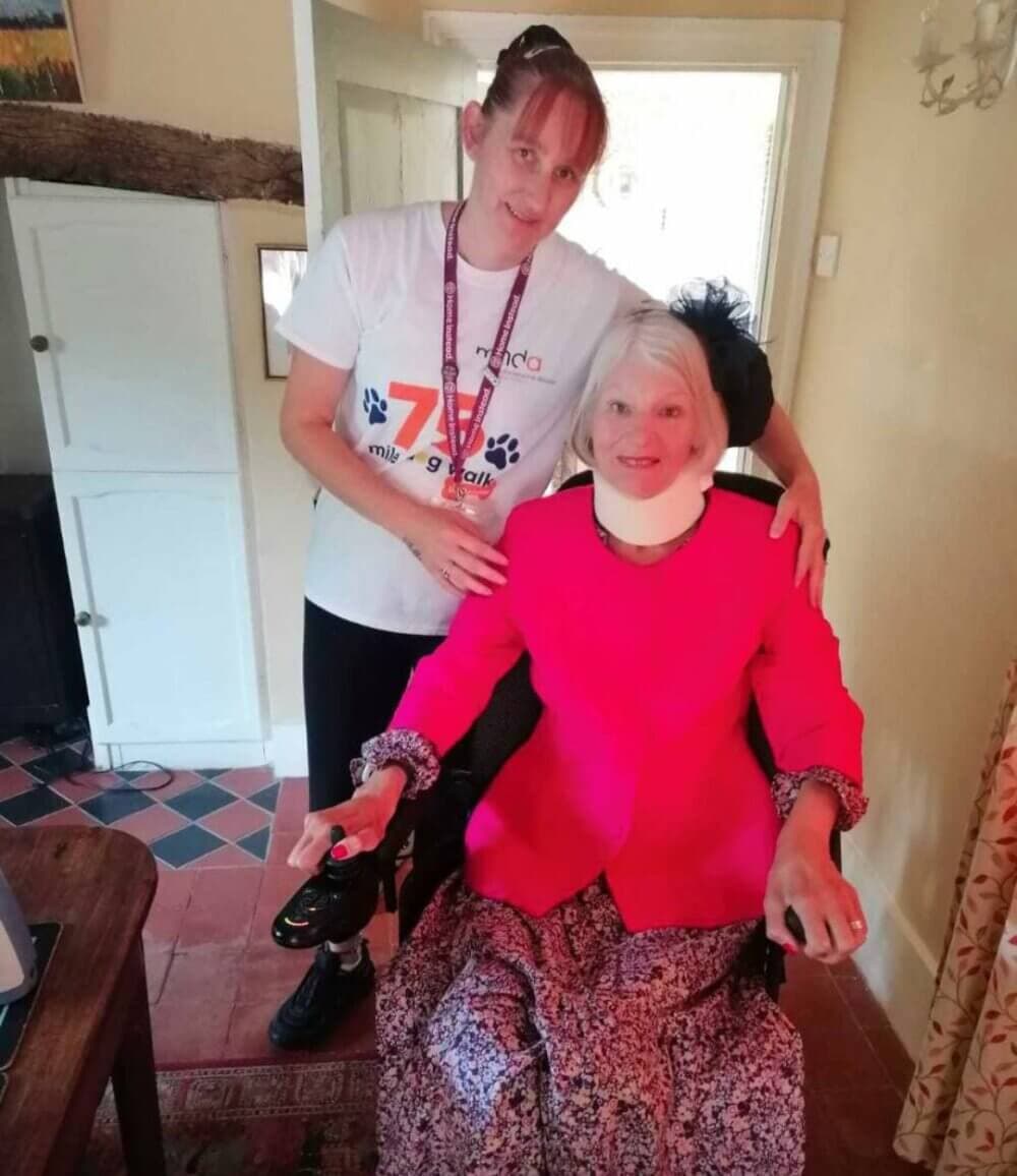 Two women, one standing in a volunteer shirt and the other in a bright pink outfit, smiling in a cozy home setting. - Home Instead