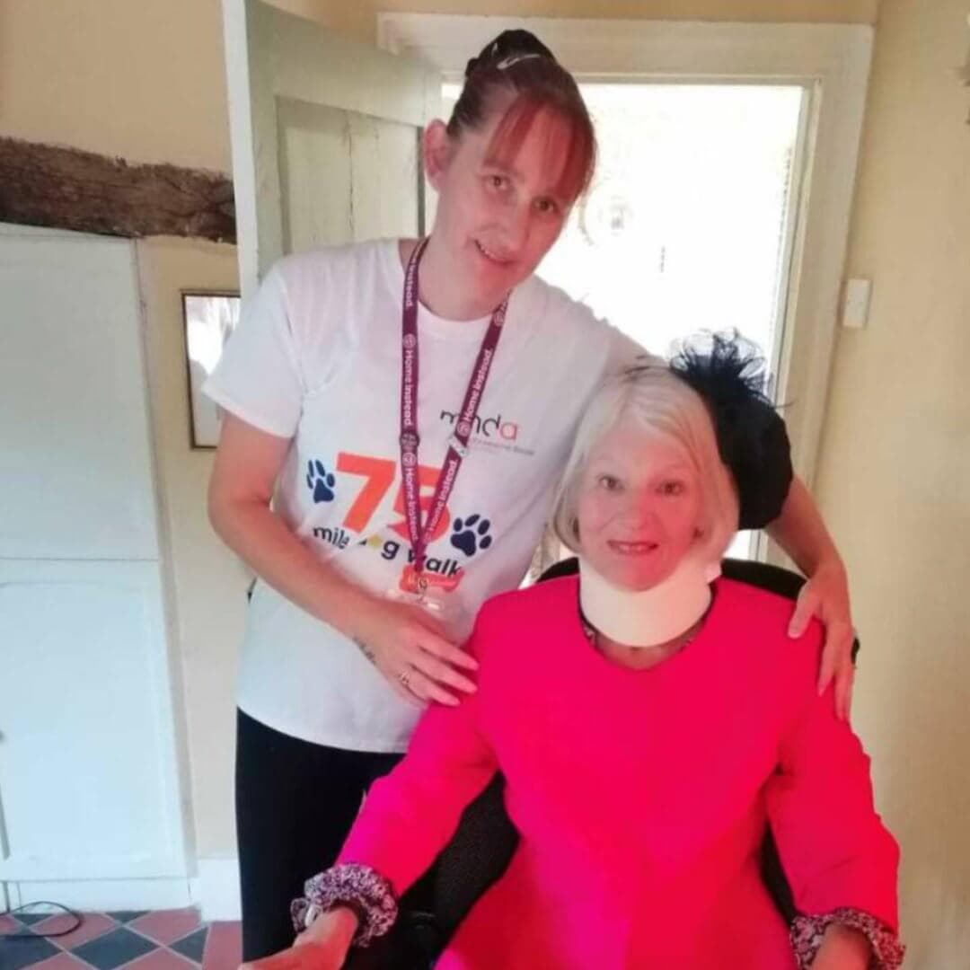 Two women, one standing in a white shirt and one seated in a bright pink outfit with a neck brace, smiling indoors. - Home Instead