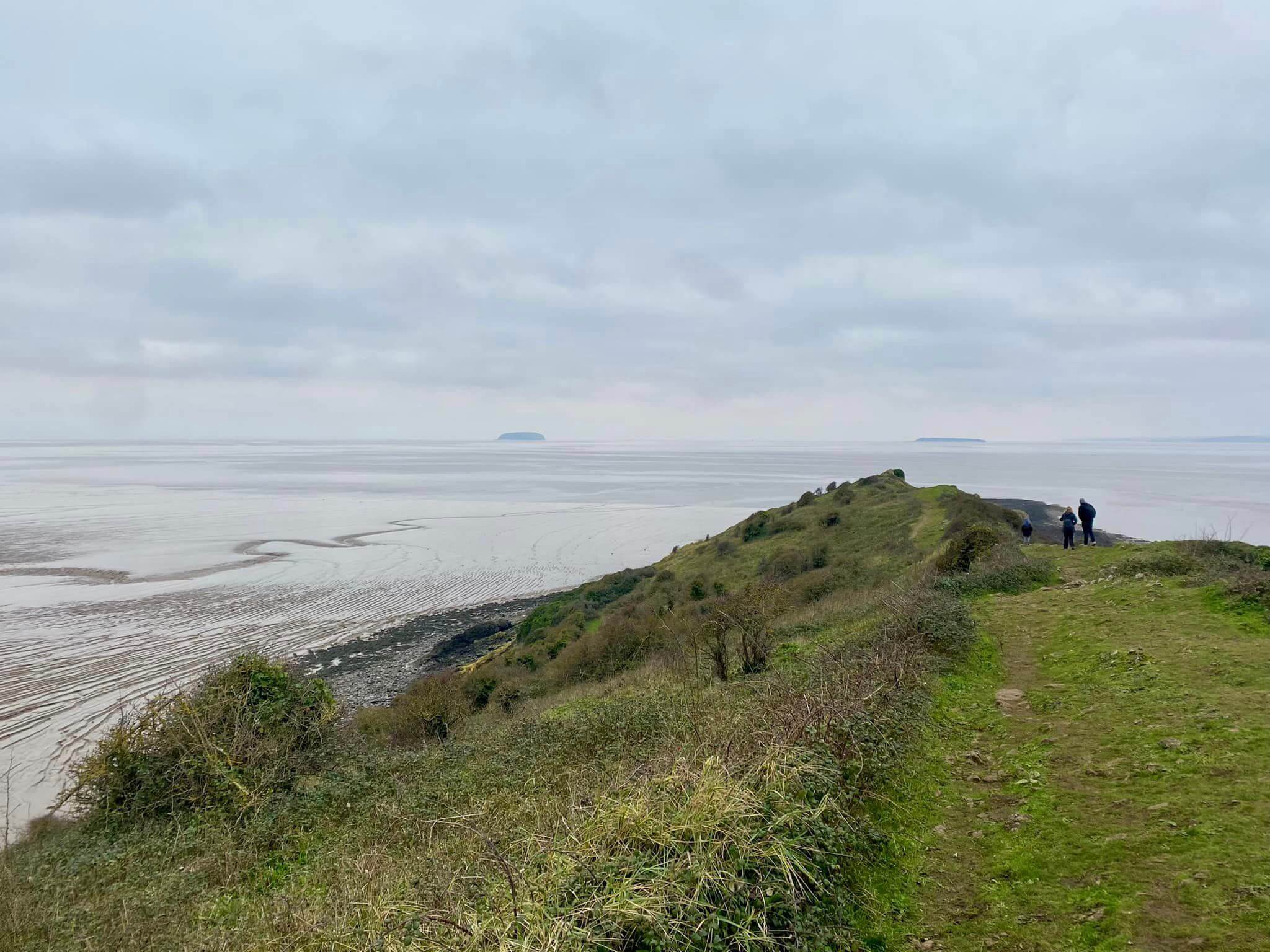 A grassy cliffside path overlooks a calm sea under a cloudy sky, with two people walking in the distance. - Home Instead