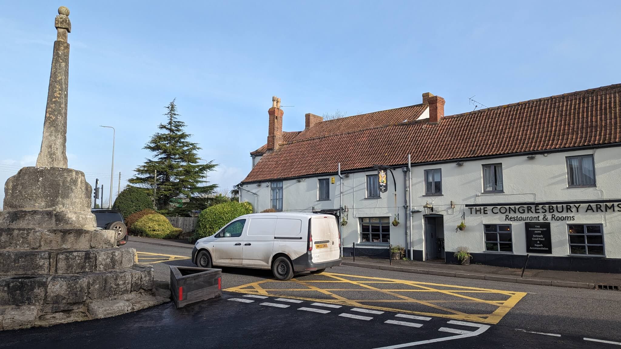 A white van is parked outside The Congresbury Arms restaurant and rooms near a stone monument on a sunny day. - Home Instead