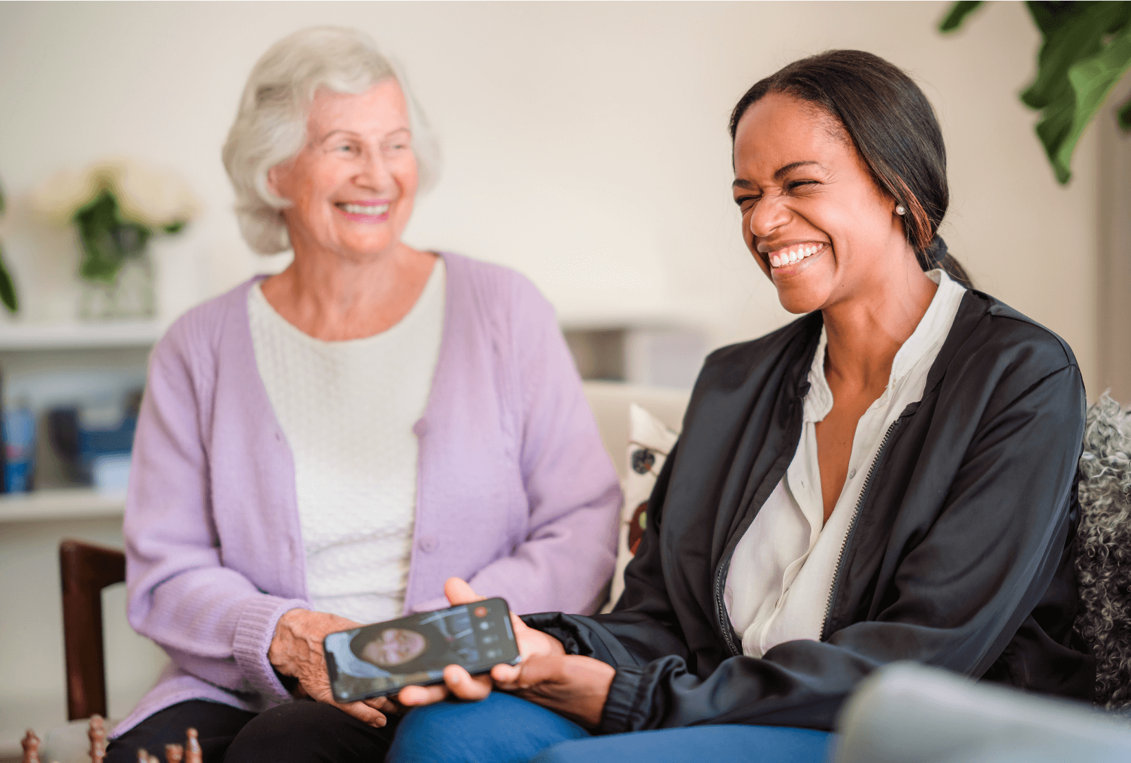 Two women, one elderly and one younger, sit together on a couch smiling; the younger woman is holding a smartphone. - Home Instead