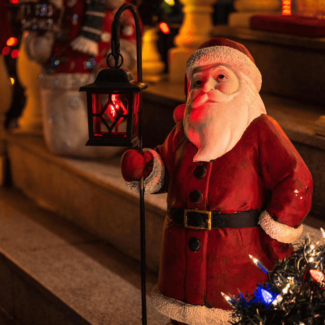 A Santa Claus figure holding a lantern, surrounded by holiday decorations with a staircase in the background. - Home Instead