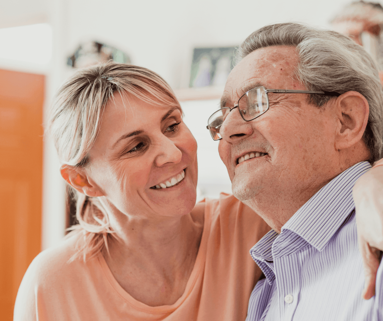 A woman smiles and embraces an elderly man wearing glasses, both sharing a warm moment indoors. - Home Instead