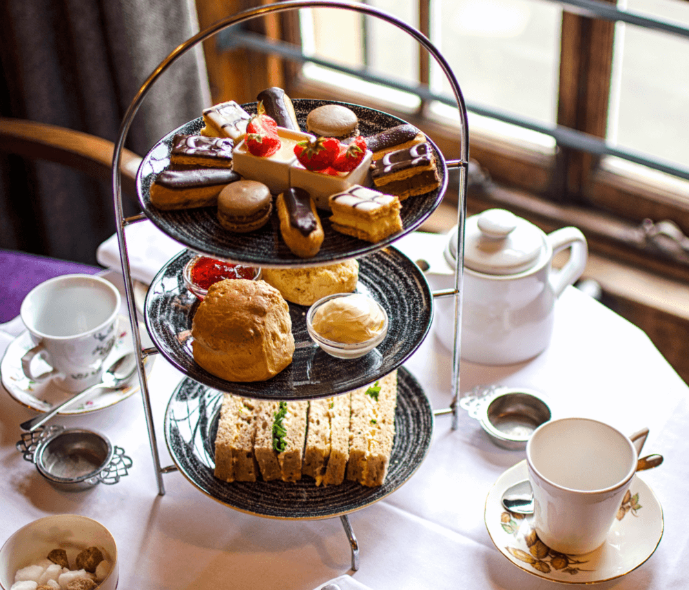 A three-tiered stand with pastries, scones, and sandwiches for afternoon tea, set on a table with teacups and teapot. - Home Instead